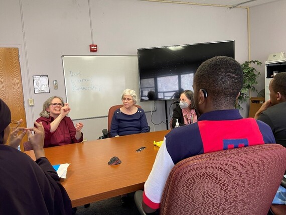 diverse group of people sit around a table in a meeting room with one woman communicating to the group using sign language