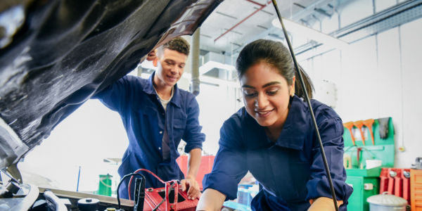 A female mechanic working underneath the hood of a car while a male mechanic observes.