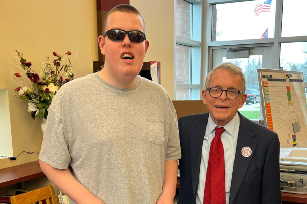 One man on the left standing and wearing sunglasses and a gray t-shirt and one man standing on the right wearing glasses and a business suit.