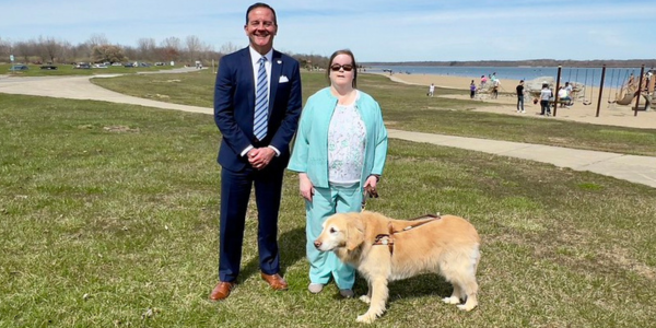 A man in business attire and woman in business attire with guide dog stand at a park.