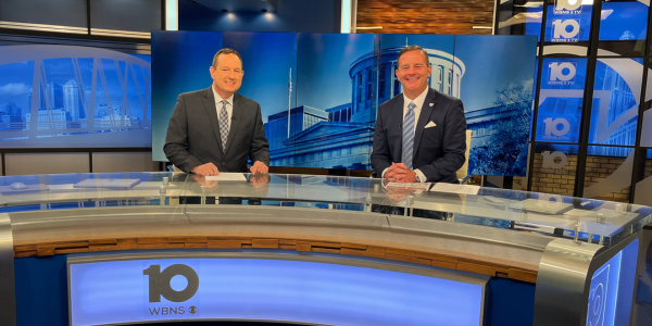 Two men wearing business attire sitting behind a news desk and smiling at the camera.