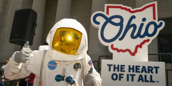 NASA astronaut giving a thumb's up gesture in front of the Ohio Heart of it All logo.