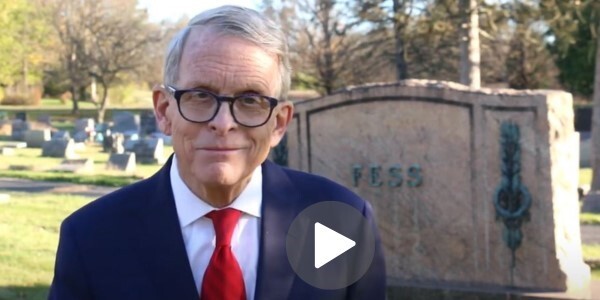 Ohio Governor Mike DeWine smiling into the camera with Senator Simeon D. Fess's headstone in the background.