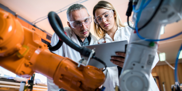 Two people dressed in personal protective equipment and lab coats review a tablet device behind a robotic device.