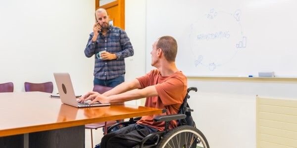 A person in a wheelchair at a desk on a computer looking over their shoulder at another person who is walking toward them.