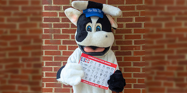 Mascot butter cow standing in front of a brick wall holding a communication board.