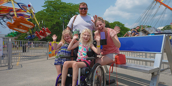Smiling family of four, including adult man, adult woman, and two young girls, posing for the camera by the Fair amusement rides.