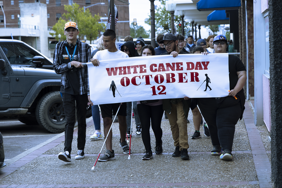 People with canes walking on sidewalk holding a sign saying White Cane Day October 12.