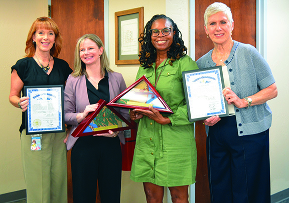 Four woman with two holding certificates and two holding Oklahoma flags in glass cases.