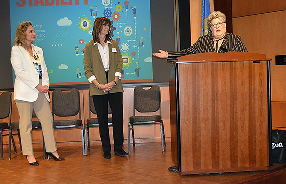 Three women on stage with one behind a podium speaking to a room of event participants.