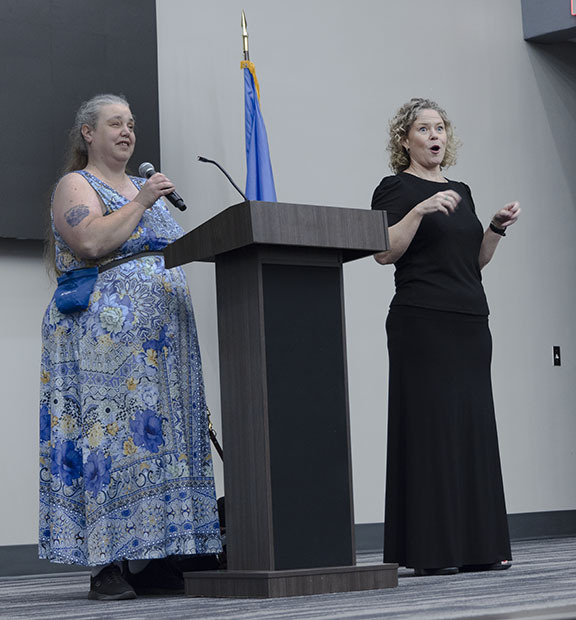 Woman holding microphone on stage and talking while a second woman uses American Sigh Language.