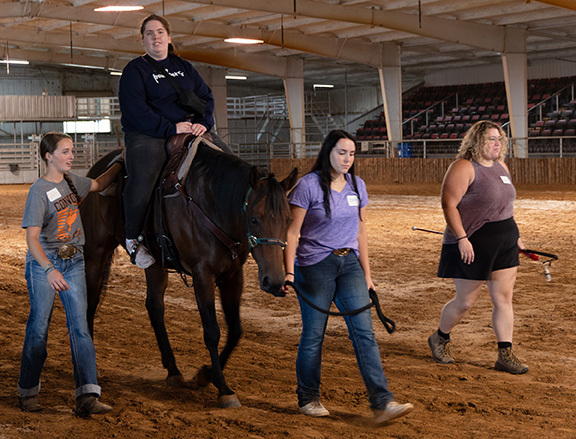 Woman riding horse with three people guiding her.