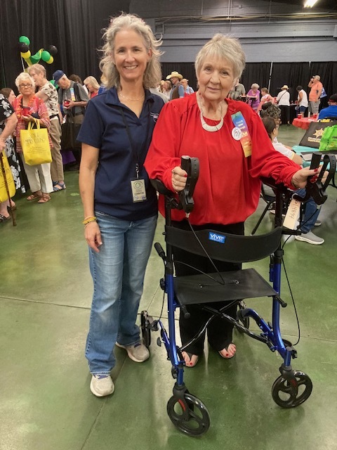 Meg and Lisa smile for a picture. Lisa is an aging light-skinned woman wearing a red shirt. She is standing with support from her 4-wheeled walker.