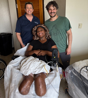 Vaniecia sits in her power wheelchair in her home. Two Device Reutilization Program staff members stand behind her. They all grin at the camera.