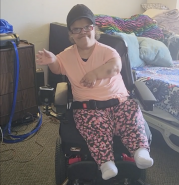 Kathrina sits in her newly acquired electric wheelchair in her bedroom. She is waving to the person taking her photo.