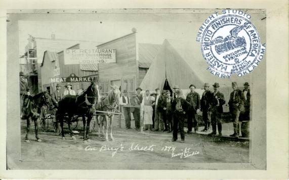 A photo from 1894 of the town of Perry. Wooden business fronts and signage with pioneers standing in foreground
