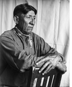a portrait of an Indigenous man seated on a chair with his hands folded