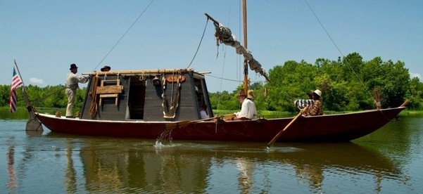 An historic keelboat with a small crew onboard, guiding the wooden vessel upriver