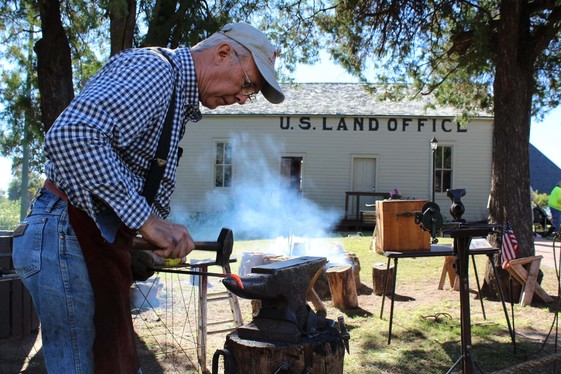 A Blacksmith working on an anvil under the shade of a tree with the historic U.S. Land Office in the background at the CSRHC in Enid