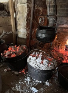 The hearth in the Fort Gibson kitchens, with embers glowing in the fireplace, a coffee pot heating at the fire, and cast iron ovens with coals on top