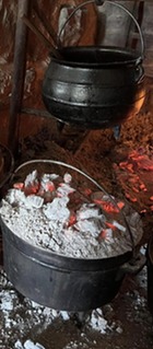 The hearth in the Fort Gibson kitchens, with embers glowing in the fireplace, a coffee pot heating at the fire, and cast iron ovens with coals on top