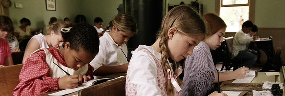 a number of schoolchildren writing in script with pen and ink at old-fashioned desks in a one-room schoolhouse.