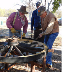 3 men gathered around a wagon wheel working to fit a metal rim on the wooden wheel