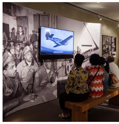 A group of people seated on a bench viewing a film about the Tuskegee Airmen, with a back drop of Black WWII soldiers