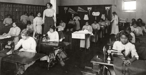 A historical photograph of a sewing class at the Chilocco Indian School. Three rows of girls working at sewing stations, some with machines