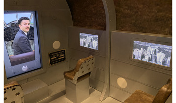 A photograph of a plane interior exhibit installation featuring a pilot and 2 window plane seats with an image of downtown Tulsa