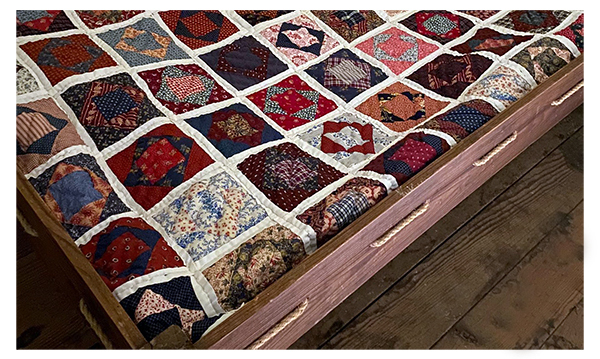 The corner of a bed located at the Hunter's Home. A potholder quilt made of multiple colors is laying on top.