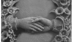 A photograph of the details of a gravestone marker with the bas relief carving of two hands touching, surrounded by flowers
