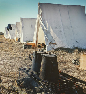 A photo taken at the Battle of Honey Springs Reenactments depicting a row of canvas tents and coffee brewing over a fire in the foreground