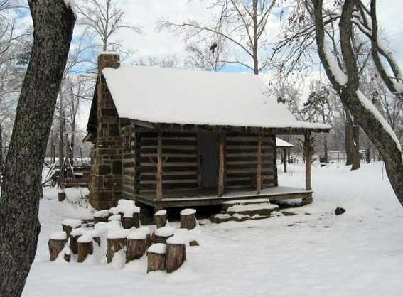 A log cabin with hand-hewn logs and a small porch after a snow storm. Snow has accumulated on the roof and surrounding grounds.