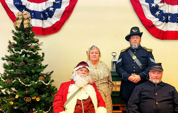 Two Civil War Union Soldier reenactors, a woman in period clothing, and Santa sit in chairs by a Christmas tree