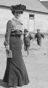 Women standing at the fairgrounds in long dresses as two boys run past. A large tent set up for carnival day Tahlequah, 1900-1902.