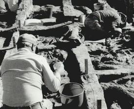 Archaeologists kneeling as they work with small tools and buckets at an archaeological dig site. 