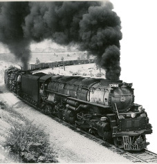 A steam engine rounding a turn with a head of steam that follows the multi-car train