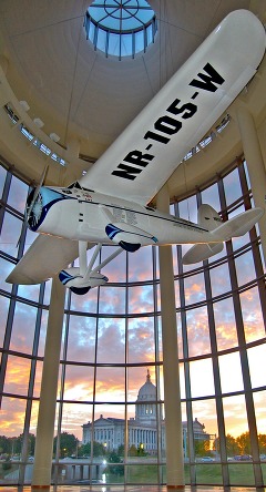Looking up at the Winnie Mae replica airplane with a sunset behind the windows of the Devon Great Hall at the OHCM.