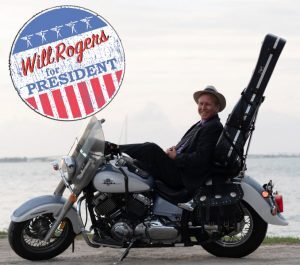 Steve McAllister leaning back on a parked motorcycle with a guitar strapped to the back. A Will Rogers for President seal.