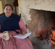 Jessica Pruitt dressed in 19th-century clothing sits by a fire reading a letter about provisions needed at Fort Towson