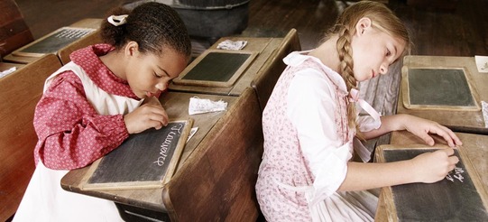 Female students in period clothing write in chalk on slates at the Rose Hill School house. The activities mimic those of 1910 school lessons.