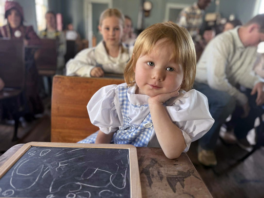 A youngster sitting at an old-fashioned shcool desk with a slate, wearing a gingham dress with her head resting on her left hand