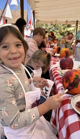 A girl painting a pumpkin wearing an apron. The table where she is working is full of parents and children decorating pumpkins.