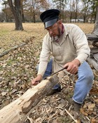 Dave Fowler dressed in period farm clothing planes the bark from a medium size tree branch in preparation for woodworking.