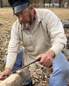 Dave Fowler dressed in period farm clothing planes the bark from a medium size tree branch in preparation for woodworking.