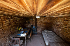 The interior of the dugout in Altus, depicting a sparse interior with a small table, 2 chairs and a single bed, and wood burner