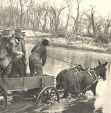 A  historic image of a wagon full of men fording a river in with two horses pulling the open wagon