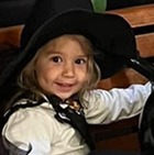 A little girl dressed as a witch for Halloween sits on a bench with balloons and Halloween decorations all around her