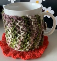 A hand-knitted mug cozy wraps a cup. The mug cozy is made up of purple, green, pink and ivory yarn.